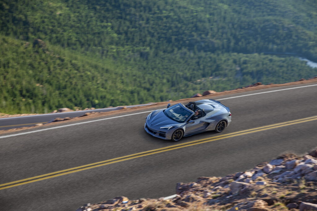 Overhead front 3/4 view of a 2024 Corvette E-Ray 3LZ convertible in Sea Wolf Gray Tricoat driving on a mountain road. Pre-production model shown. Actual production model may vary. Model year 2024 Corvette E-Ray available 2023.