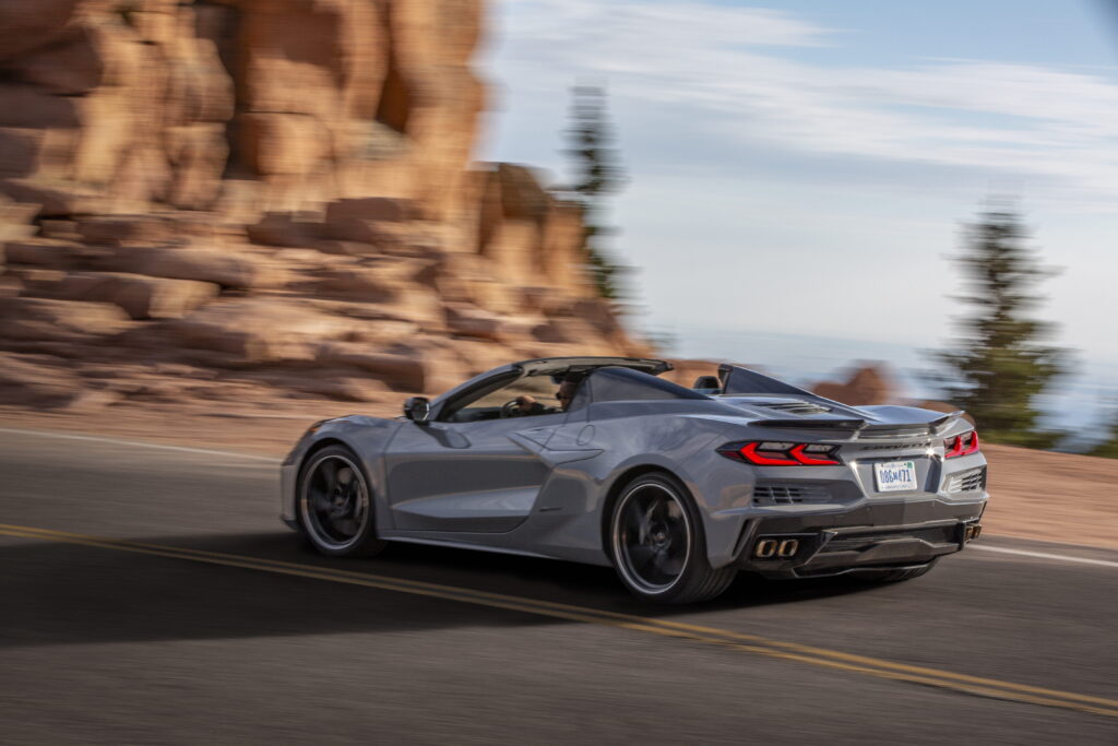 Rear 3/4 view of a 2024 Corvette E-Ray 3LZ convertible in Sea Wolf Gray Tricoat driving on a mountain road. Pre-production model shown. Actual production model may vary. Model year 2024 Corvette E-Ray available 2023.